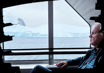 Professor Eric Eckert sitting near a window on the ship with a view of the snow covered land of Antarctica