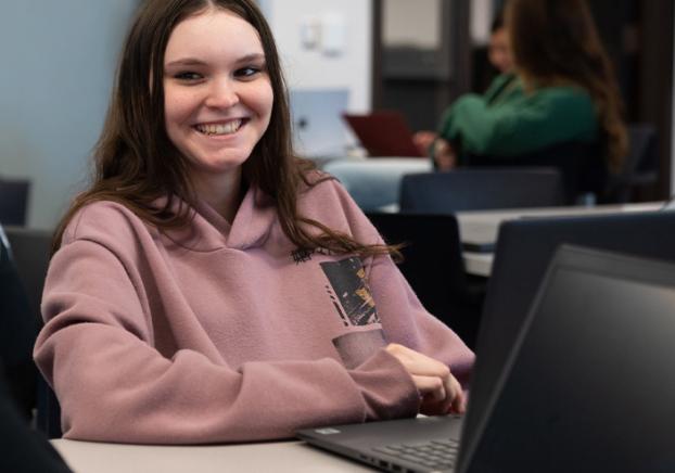 In a classroom, a young college student smiles while sitting in front of her laptop.