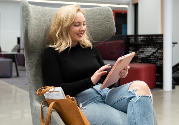 A young college female sits in a chair and smiles while viewing her digital tablet. 