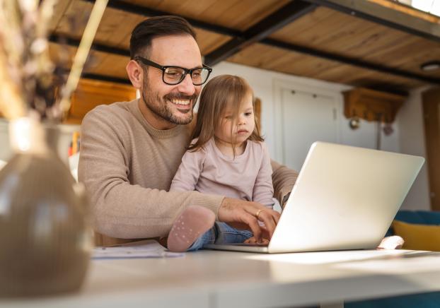 A father working on his laptop with his toddler daughter sitting in front of him
